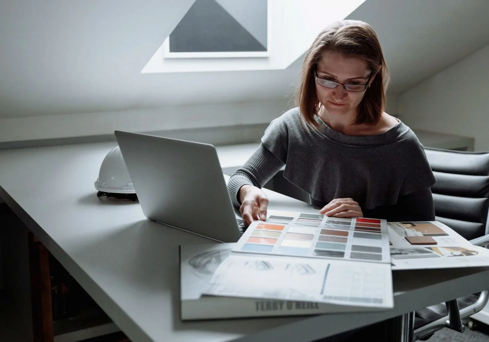Businesswoman analyzing color palettes at her desk with a laptop and a hard hat nearby, in a modern office.